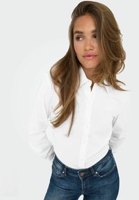 Camicia bianca a maniche lunghe con bottoni, con colletto classico, abbinata a jeans aderenti blu scuro. Tessuto liscio con texture minima.