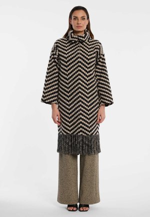 Woman wearing a long black and beige chevron-patterned coat with fringe at the hem, wide-legged beige pants, and black open-toe shoes.