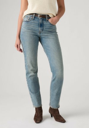Kvinde iført lyseblå højtaljede jeans med brun bælte, cremefarvet striktop og brune ruskindsstøvler til anklerne, stående med den ene hånd i lommen.