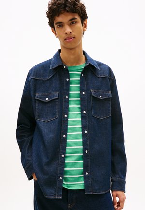 Giovane uomo che indossa una giacca di jeans blu scuro sopra una camicia a righe verdi e bianche, in piedi con le mani in tasca davanti a uno sfondo semplice.