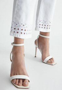 Sandalias blancas de tacón alto con un diseño de punta abierta, tiras delgadas en el tobillo y una punta cuadrada. Combinadas con pantalones blancos que presentan un borde de ojales en el dobladillo.