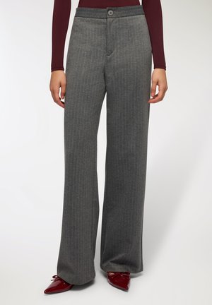 Pantaloni a gamba larga grigio a righe con una texture liscia, dotati di chiusura frontale con bottone e vita. Indossati con scarpe rosse a punta.