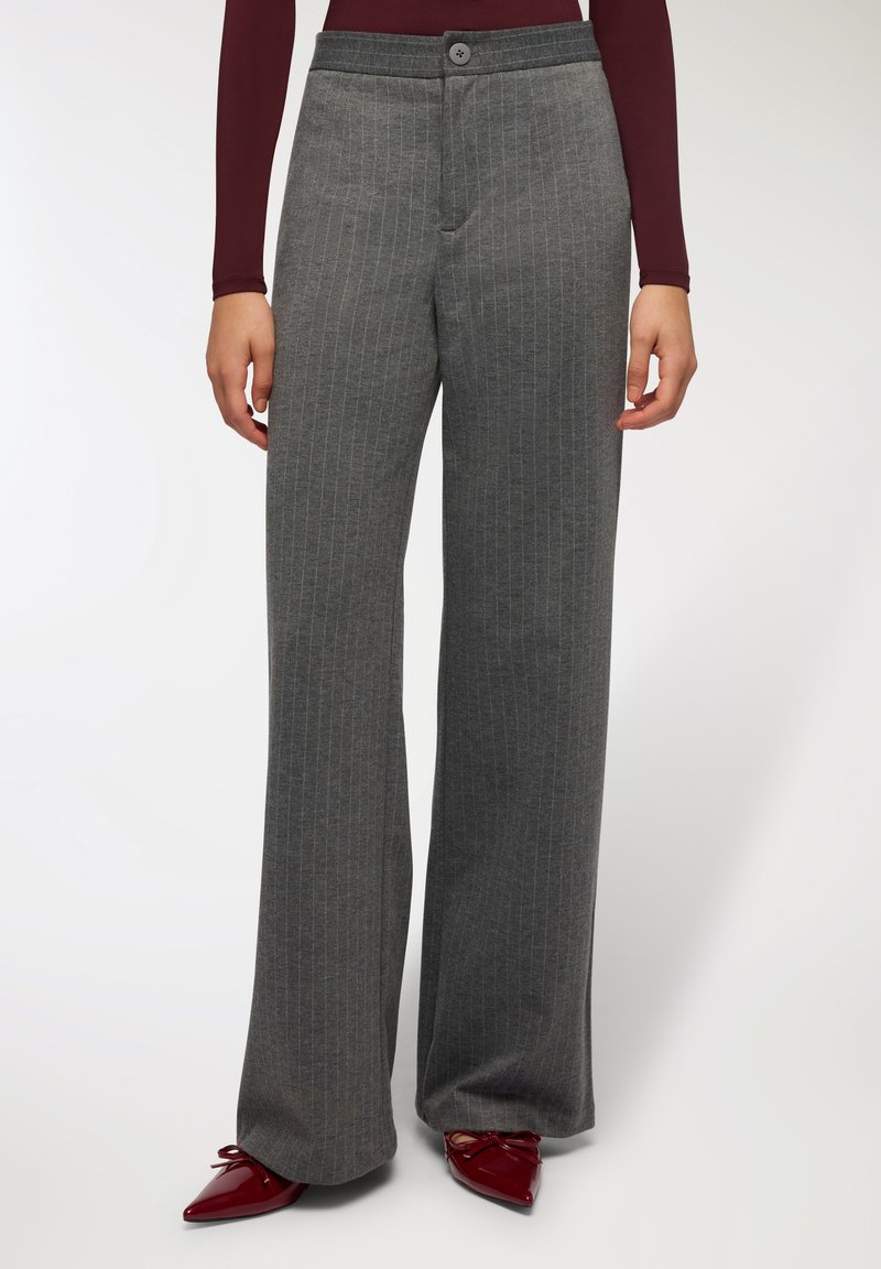 Szerokie spodnie w szary prążek o gładkiej teksturze, z zapięciem na guzik z przodu i pasem. Noszone z czerwonymi, spiczastymi butami.