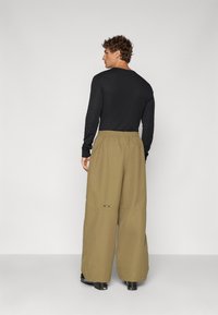 Svart langermet skjorte og khakifargede vide bukser med strikk i midjen, med en brodert logo nær kanten. Glatt teksturfinnish.