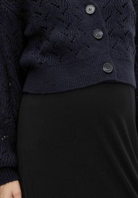 Mörkblå stickad kofta med kabelmönster och svarta knappar, kombinerad med en svart kjol, som visar en närbild av midjan och handen.