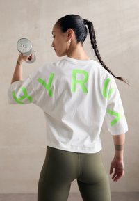 Biely skrátený tričko s odvážnymi limetkovozelenými písmenami "HYRG" na chrbte. Modelka nosí olivové legíny a drží 6kg činku.