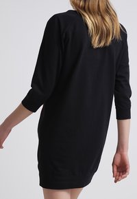 Czarna sukienka z rękawami do trzech czwartej, miękka tkanina, nieco luźny krój i zaokrąglony dół. Minimalistyczny design, bez widocznych wzorów czy akcentów.