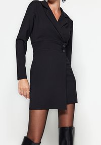 Černé sako šaty s dlouhými rukávy, vypasovaným pasem a dvouřadým zapínáním. Hladká tkanina a elegantní design s délkou po stehna.