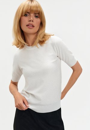 Biały sweter z krótkim rękawem, wykonany z dzianiny z delikatnym połyskiem, z prążkowanym okrągłym dekoltem i dopasowaną talią. Noszony z czarnymi spodniami.