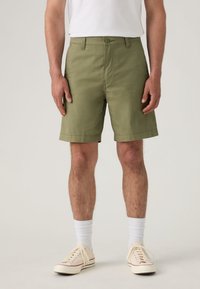 Shorts en coton vert olive à coupe droite, avec poches avant et fermeture par bouton. Portés avec des baskets crème et des chaussettes blanches montantes.
