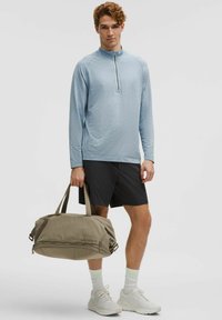 Ljusblå långärmad topp med dragkedja, svarta shorts och en beige duffelväska som hålls i handen. Texturerat tyg, atletisk design, neutrala färger.