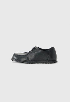Sapatinho de couro preto com biqueira arredondada, lados baixos e acabamento texturizado. Apresenta um sistema de atacadores simples e uma sola de borracha flexível.