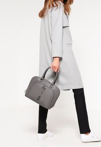 Borsa strutturata grigia con cerniera, manici corti, un accento a nappa e una texture simile alla pelle liscia, abbinata a pantaloni neri e sneakers bianche.