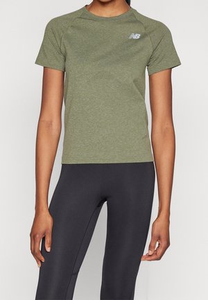 Persoon draagt een olijfgroen sportshirt met korte mouwen met New Balance-logo en zwarte aansluitende legging, staand tegen een effen achtergrond.