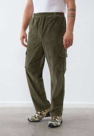 Homme portant un pantalon cargo en velours côtelé vert olive avec taille élastique et chaussures de trail camouflage vert, debout sur un sol gris.