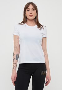 T-shirt bianca a maniche corte con scollo rotondo, realizzata in tessuto morbido, con una silhouette aderente. Indossata con leggings neri.