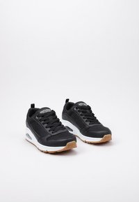 Zapatillas deportivas negras con parte superior de malla y sintético, diseño con cordones, suela con amortiguación, media suela blanca y suela de goma con un patrón texturizado.