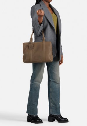 Bruine leren tote bag met een gestructureerde afwerking, dubbele bovenhandvatten en logodetail. Het model draagt een grijze blazer over een groene top en lichte jeans.