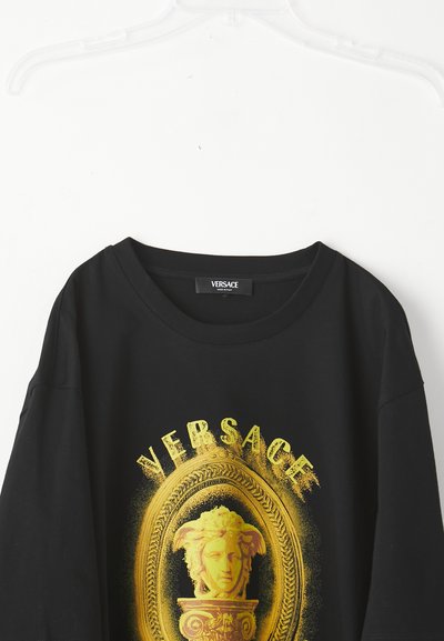 Black Versace T-shirt on a hanger with a large yellow Medusa head and decorative frame graphic on the front.