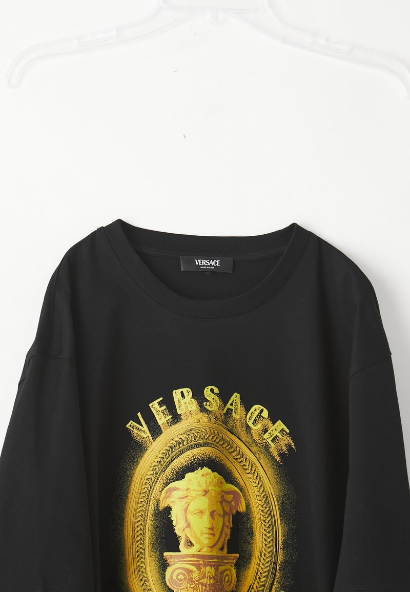 Zwarte Versace T-shirt aan een hanger met een groot gele Medusa-kop en een decoratief kader grafiek op de voorkant.