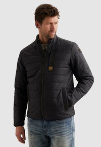 Muž v čiernej prešívanej bunde so zipsom, rukami v vreckách, pozerajúci sa do strany, oblečený v svetlomodrých džínsoch na jednoduchom pozadí.