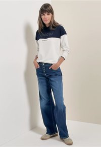 Sudadera blanca y navy con el texto "UNIQUE", combinada con unos vaqueros azules holgados. El modelo lleva zapatos beige, con las manos en los bolsillos, de pie contra una pared clara.