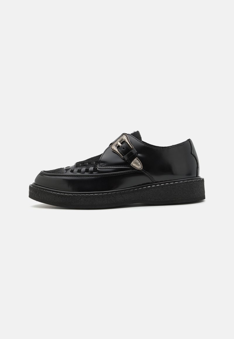 Des chaussures en cuir noir avec une finition brillante, une sangle texturée et une boucle décorative en argent. Elles présentent une semelle épaisse et texturée pour une meilleure adhérence.