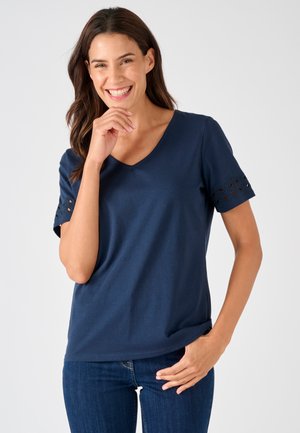 Donna sorridente che indossa una T-shirt blu scuro con scollo a V e maniche corte con pizzo, e jeans blu, in posa con una mano vicino al viso e una in tasca.