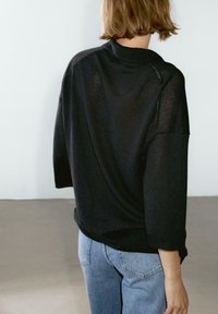 Person with short hair wearing a loose black sweater with three-quarter sleeves and light blue jeans, standing against a plain light background.