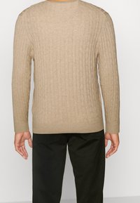 Osoba v béžovém svetr s copánkovým vzorem a dlouhými rukávy a černých kalhotách, zobrazena zezadu na jednobarevném světlém pozadí.