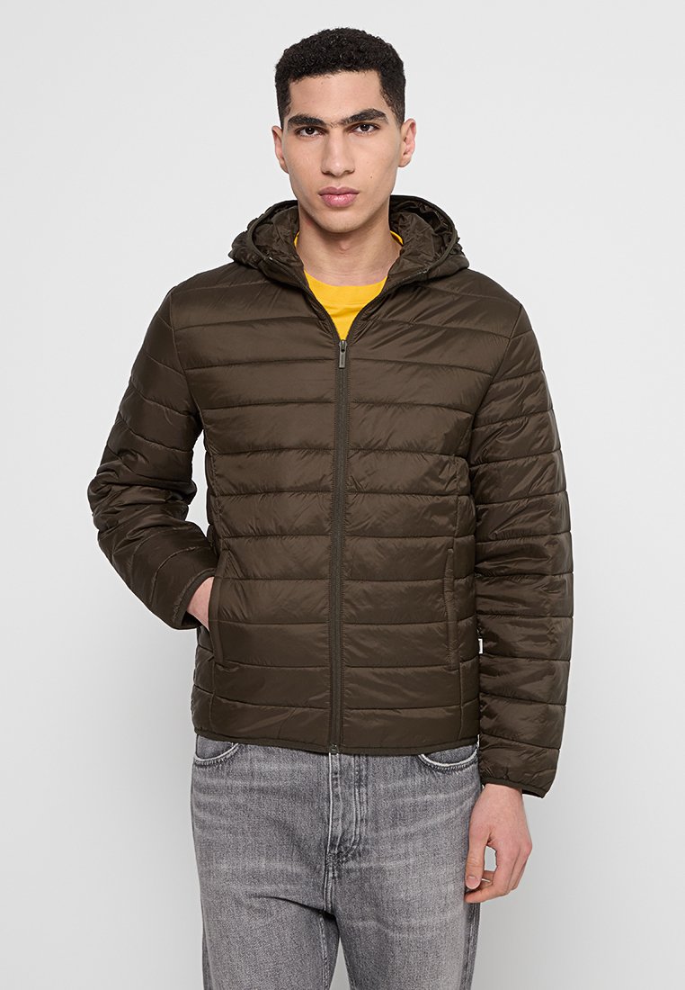 Veste rembourrée marron avec fermeture éclair sur le devant, capuche et poches latérales. Présente un design matelassé et une texture lisse et brillante. Portée par-dessus une chemise jaune.