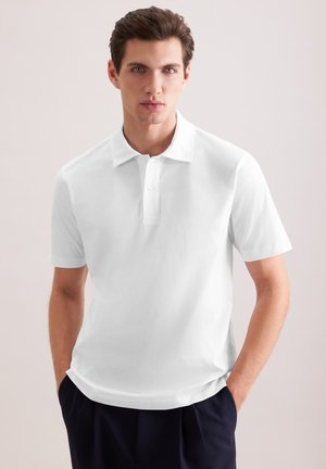 Giovane uomo con capelli corti castani che indossa una polo bianca a maniche corte e pantaloni scuri con le mani in tasca su uno sfondo semplice.