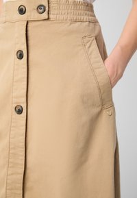 Jupe midi beige en coton avec un devant boutonné, des poches latérales, une taille élastique et un petit détail doré.