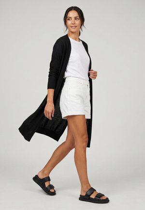 Femme marchant, portant un long cardigan noir, un t-shirt blanc, un short blanc et des sandales noires sur un fond uni clair.