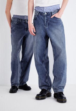 Jasnoniebieskie jeansy z denimu o luźnym kroju, z paskiem z tkaniny w paski, przednimi kieszeniami i zużytym wykończeniem. Czarne buty dopełniają wygląd.