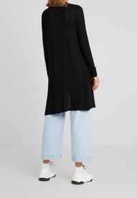 Black long-sleeve top with a relaxed fit, featuring a split hem and lightweight fabric, paired with light blue wide-leg jeans and white sneakers.