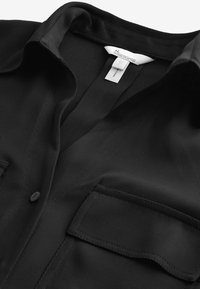 Černá košile s límečkem vyrobená z žebrované tkaniny s nenápadným leskem. Má vertikální pruhovaný vzor a dvě přední kapsy.