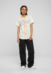 Maglietta da baseball bianca con righe in rilievo, logo in corsivo nero, maniche corte, abbinata a pantaloni cargo neri e scarpe da ginnastica bianche.