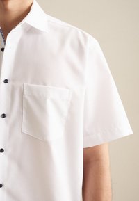 Camicia bianca a maniche corte con tasca, dotata di bottoni neri e rivestimento a collare a motivo. Tessuto leggero e liscio.