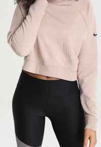 Sweat-shirt court rose clair avec logo texturé, associé à des leggings noirs taille haute avec des accents gris sur le bas des jambes.