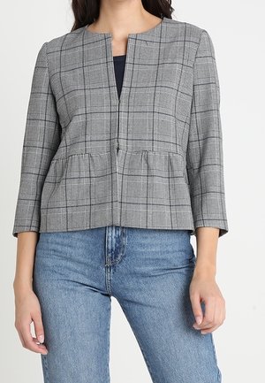 Vrouw die een grijs geruit jack met rits draagt, met driekwart mouwen en een blauwe spijkerbroek, staand tegen een effen witte achtergrond.