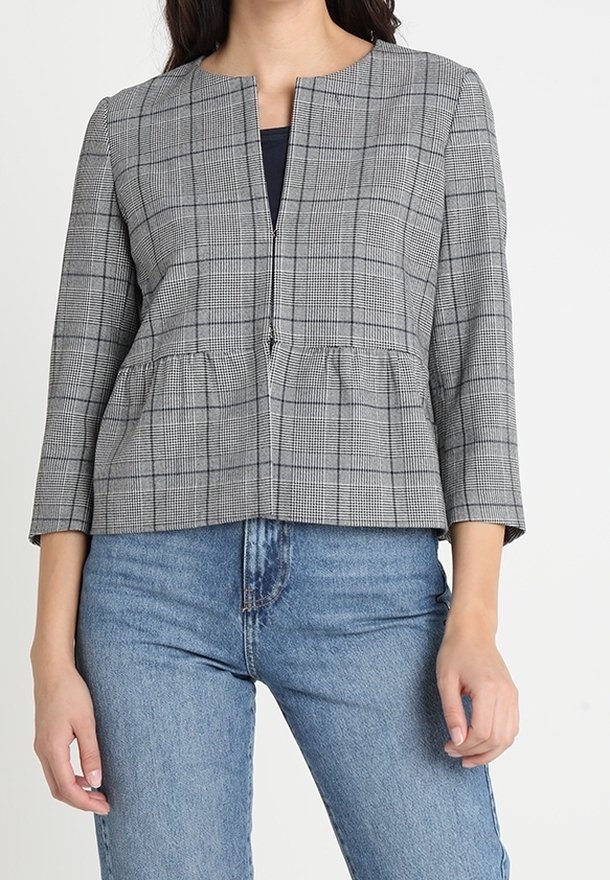 Femme portant une veste zippée grise à carreaux avec manches trois-quarts et un jean bleu, debout devant un fond blanc uni.