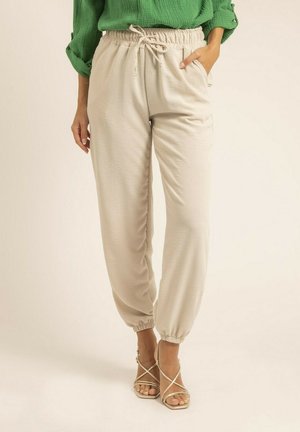 Pantalons de jogging beige légers avec une taille élastique et un cordon de serrage, dotés de poches latérales et de poignets fuselés, associés à des sandales dorées à lanières.