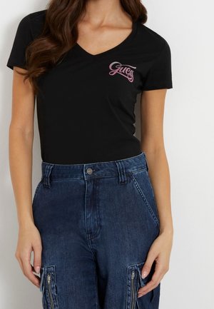 Kobieta w czarnym T-shircie z dekoltem w serek z różowym logo "Guess" i wysokim stanem niebieskich dżinsów z kieszeniami na zamki na udach.