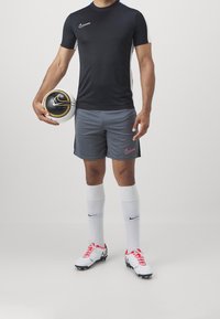 Tenue de sport Nike comprenant un t-shirt noir à manches courtes et un short gris. Comprend des chaussettes blanches et des chaussures à crampons blanches et rouges. Tient un ballon de football noir et doré.