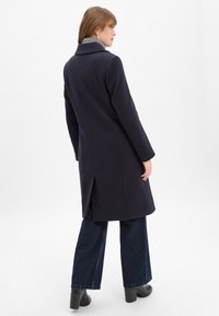 Marineblauer Mantel mit taillierter Silhouette, langen Ärmeln und einem Schlitz hinten. Kombiniert mit weiten, dunklen Jeans und Absatzstiefeln.