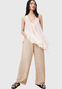 Vloeiende crème blouse met plooien en ruches, gecombineerd met losse beige broeken. Model draagt zwarte sandalen; minimale accessoires aanwezig.