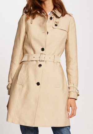 Woman wearing beige belted trench coat with black buttons and buckled wrist straps, paired with blue jeans.