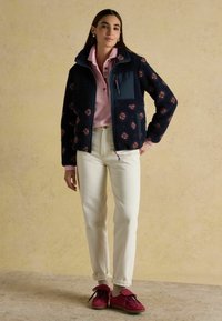 Donkerblauw fleecejack met bloemmotieven, roze shirt, witte spijkerbroek en bordeauxrode sneakers. Het jack heeft een ritssluiting en een zachte textuur.