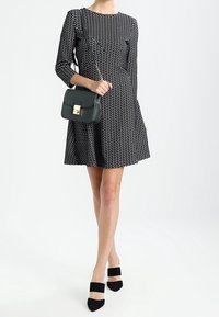 Robe à pois noirs et blancs avec manches longues, jupe évasée et une poche avant. Associée à un petit sac à bandoulière vert et des chaussures noires à talons carrés.
